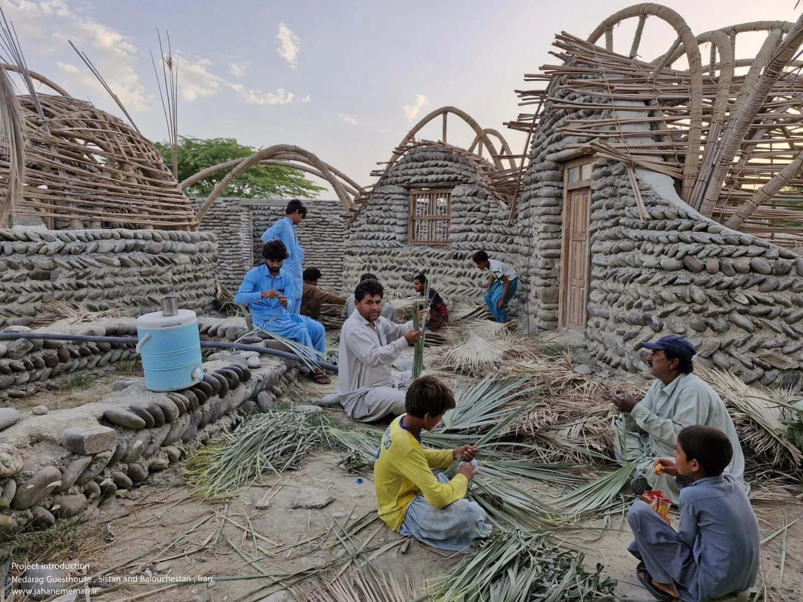 نمای خارجی و داخلی مهمانسرای ندارگ در روستای کهنانیکش، سیستان و بلوچستان، که ساختار نیمه‌باز، سقف چتری‌شکل با خرپاهای دست‌ساز و دیوارهای ضخیم گلی آن بر اساس بازتفسیر معماری بومی «کپر» و با هدف خنک‌سازی طبیعی و مشارکت جمعی اهالی طراحی و اجرا شده است.