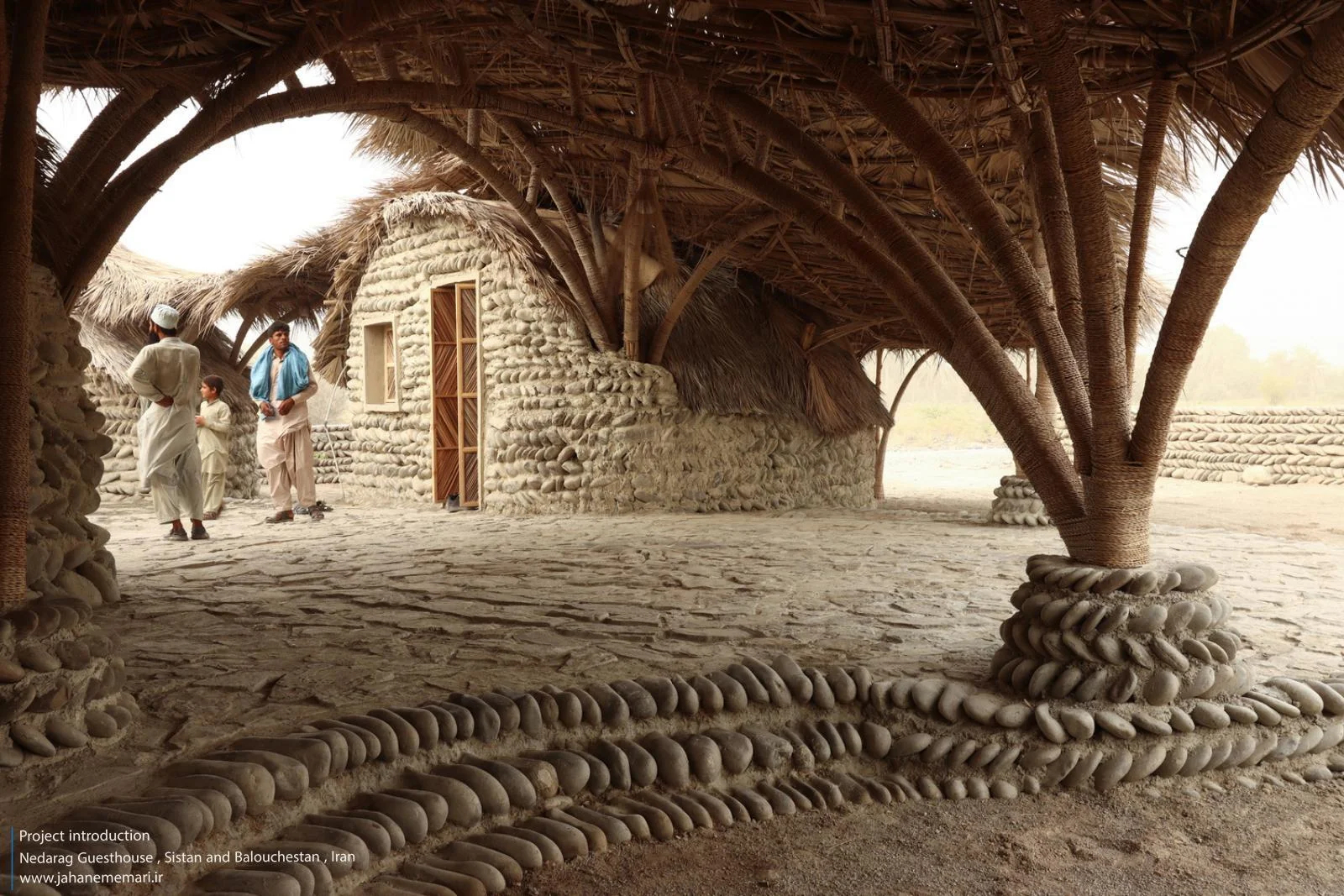 نمای خارجی و داخلی مهمانسرای ندارگ در روستای کهنانیکش، سیستان و بلوچستان، که ساختار نیمه‌باز، سقف چتری‌شکل با خرپاهای دست‌ساز و دیوارهای ضخیم گلی آن بر اساس بازتفسیر معماری بومی «کپر» و با هدف خنک‌سازی طبیعی و مشارکت جمعی اهالی طراحی و اجرا شده است.
